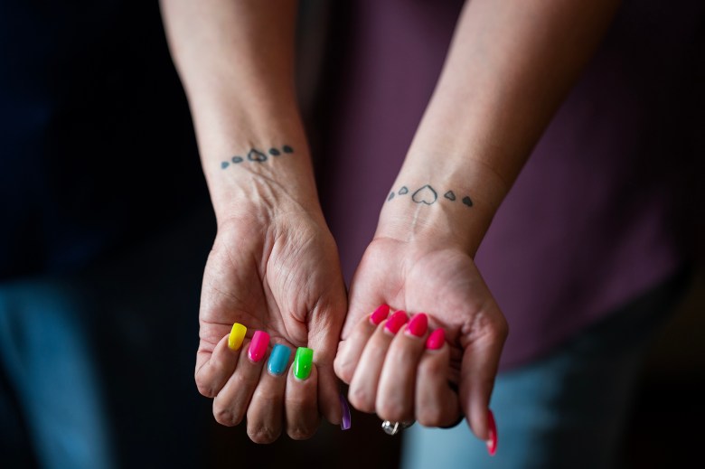 Two hands extended forward, showing wrists with matching heart tattoos in a row, with the central heart being outlined and flanked by smaller, filled hearts. The nails are painted in vibrant, multicolored shades, with one hand featuring yellow, pink, green, blue, and purple nails, and the other sporting bright pink nails.
