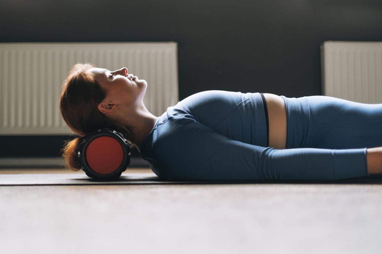 young red haired woman in blue sportswear with pilates roller under neck doing exercises in the studio