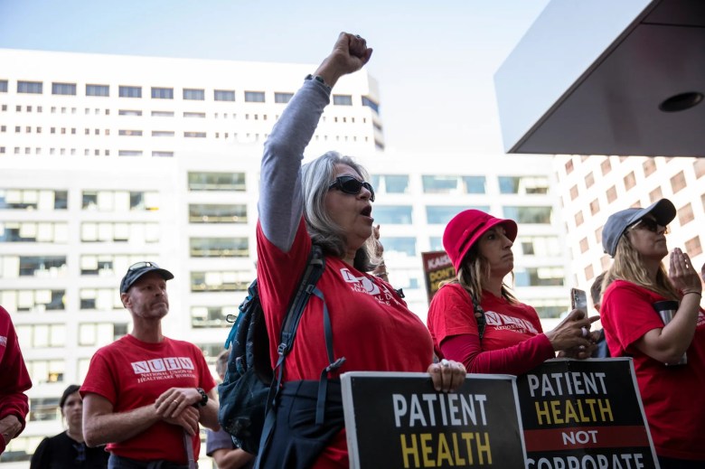 A group of protesters, mostly wearing red shirts with "NUHW" logos, during a demonstration. A person with gray hair raises a fist in the air, alongside signs with advocacy messages, with a white building in the background. Courtesy CalMatters