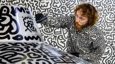 Getty Images Mr Doodle to the right of screen wearing a shirt covered in his own doodles, with a doodle background and drawing on a structure in the foreground with a big black marker pen