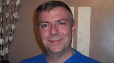PA Real Life Brian Place, who has grey, short hair, smiling at the camera. He's wearing a blue t-shirt.