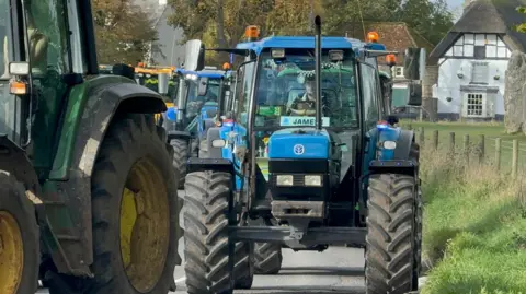 Wiltshire Agricultural Preservation Group A close up of a blue tractor driven by a young man with a licence plate reading "James" in the cab window. It is part of a line of tractors passing through a country village. 