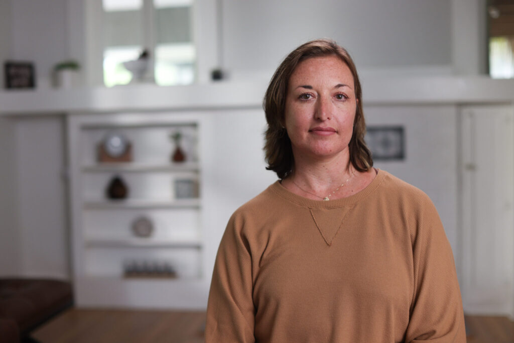 A woman in a brown long-sleeved shirt in a room with shelves visible in the background