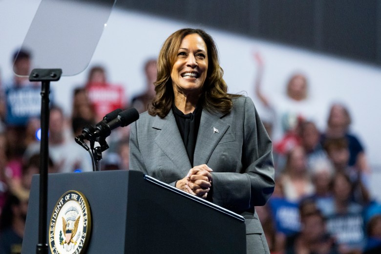 Kamala Harris smiles while standing behind a podium with two microphones and a presidential seal and her hands clasped.