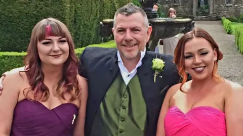 PA Real Life Brian Place wearing a suit and green waistcoat standing with his arms around his two daughters. The woman on the left has long, curly, brown and pink hair and is wearing a purple dress. The woman on the right has brown and orange curly hair and is wearing a pink dress. 