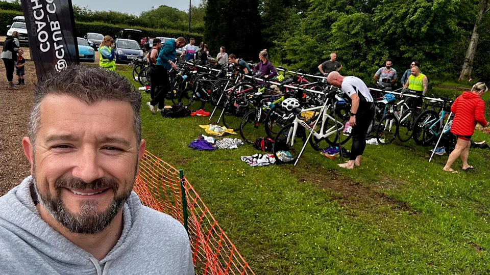 A man with short black and grey hair wearing a grey hoodie takes a selfie with people stood behind him in exercise clothing and racks of bikes. 
