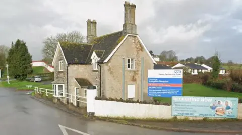 Google Google Street View image of the entrance to the Langdon Hospital site in Dawlish, Devon. A house which is part of the site is next to the junction of the road where the entrance is. A blue, white and red sign with the hospital's name on it along with other directional information is next to a white stone wall. A sign with a recruitment advert on it is also on the wall.