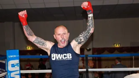 Robert Edwards A man stands in a boxing ring with both his arms raised and his mouth wide open as he celebrates. He has red wraps around his hands and wrists, tattoos covering his arms and is wearing a blue tank top. 
