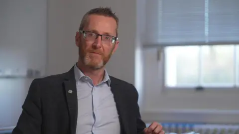 A man with short, dark hair spiked at the front. Wearing black glasses, a grey blazer and blue shirt. He is pictured against a cream wall with a window behind him.