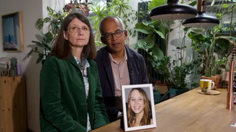 Jane and Max Figueiredo sitting at a table looking at the camera, with a framed, smiling photo of Alice, a woman with long blonde in the foreground. Jane is wearing a green blazer, glasses and has shoulder-length dark hair. Max is balding, wears glasses, and wears a dark blue blazer. There are many plants in the background. 