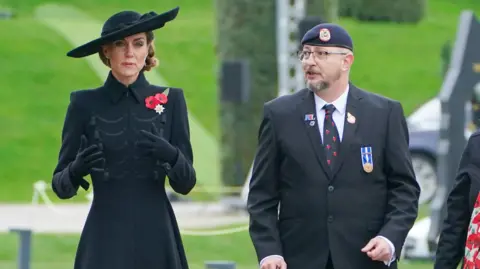 PA Media The Princess of Wales is walking beside a man in a black suit and navy beret. She is dressed in a large black hat, black gloves, a long black coat with two red poppies on her left chest and her brown hair is tied up. The man is wearing a white shirt, black tie decorated with poppies, a medal and three pin badges, one of which is shaped like a poppy. Grass, a headge and a guidance rope can be seen blurred out behind them.