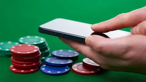 Getty Images A hand holding a black smartphone, hovering over a selection of green, red and blue poker chips