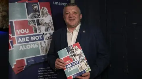 John Fairhall/BBC Nigel Seaman smiles at the camera as he holds the new book. He has short dark and grey hair and wears a navy suit jacket with a white shirt underneath. A poster board promoting the new book stands behind him.