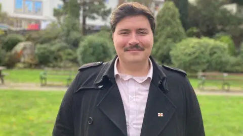 BBC Tom Rylatt pictured smiling at the camera. He has short dark brown hair and a moustache. He is wearing a black coat with a pink button down shirt.