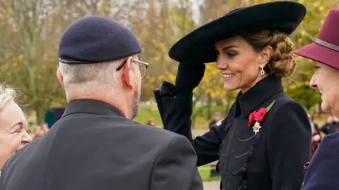 PA Media The Princess of Wales is standing in front of one man and two women. She is holding a large black hat on her head and wearing black gloves, a long black coat with two red poppies on her left chest and her brown hair is tied up. The man has his back to the camera and is wearing a black suit, navy beret and glasses. The woman to the right is wearing a maroon hat and a navy coat. The lady on the left has long blonde hair, a black coat and a scarf decorated with poppies. There are lots of blurred out trees behind them.