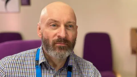 Dorian pictured in the offices of Derbyshire Healthcare NHS Foundation Trust wearing a checked shirt and an NHS lanyard.