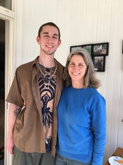 A young man and an older woman stand side by side indoors, smiling at the camera. Several framed photos hang on the white-paneled wall behind them.