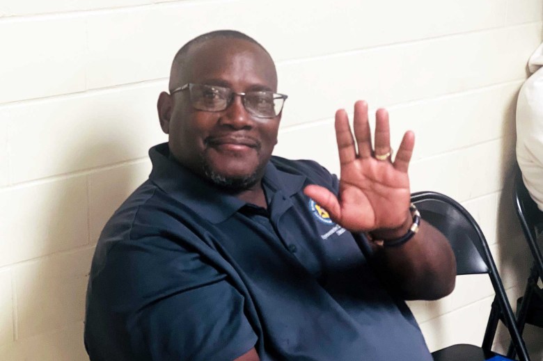 A seated man wearing glasses, waving to the photographer