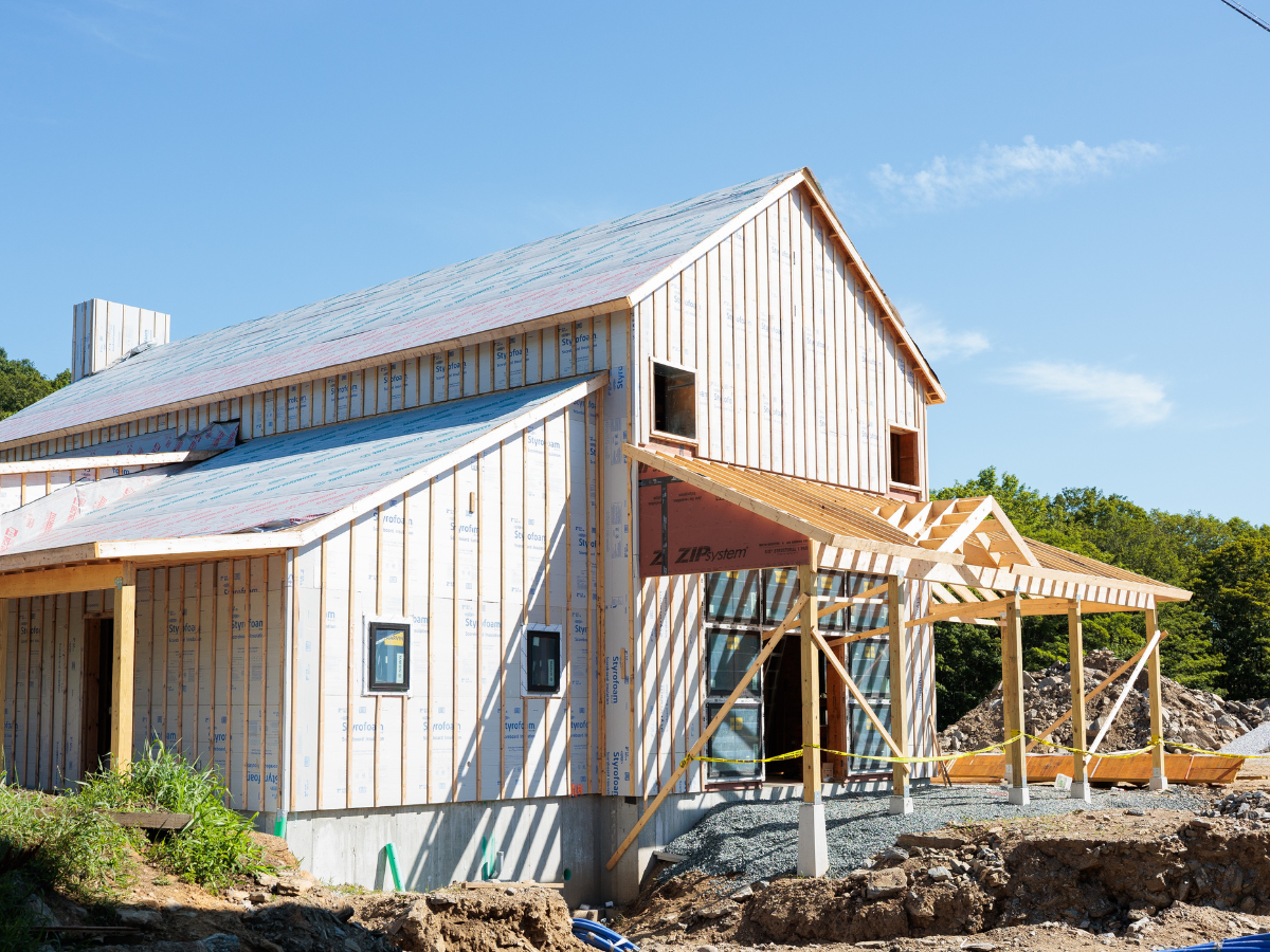 A two-story building under construction with exposed framing, insulation, and a partially built porch roof on a sunny day.