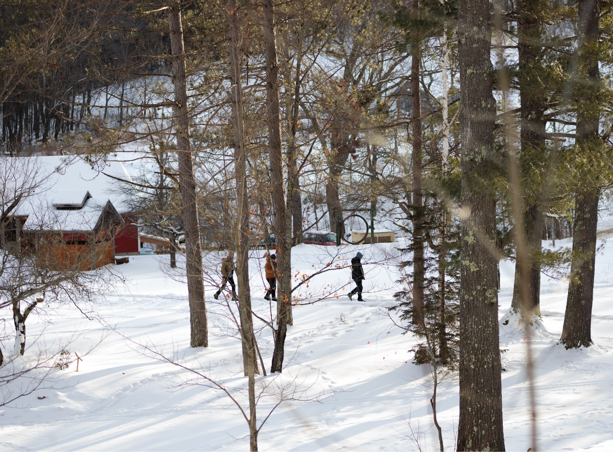 Three people walk through a snowy forested area near red buildings, with tall trees and snow covering the ground.