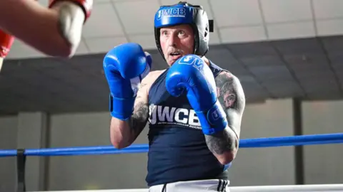 Robert Edwards A man wearing boxing attire, including a blue helmet and gloves, stands in a boxing ring with his hands raised ready for a fight. He is wearing a dark blue sleeveless shirt and there are tattoos covering both his arms.