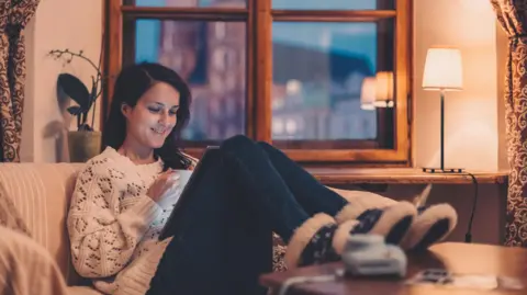 Getty Images A woman with brown hair wearing a white jumper, jeans and fluffy shoes looking at her tablet sat on the sofa. 