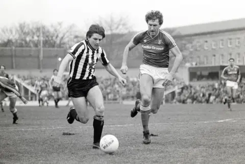 Danny Brannigan/Hulton/Getty A black and white picture of Terry Butcher, of Ipswich Town, chasing alongside Peter Beardsley of Newcastle United, who has the ball at his feet. Open terracing at St James' Park is in the background, with flats behind the ground.