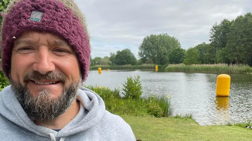 A man wearing a purple woolly hat and grey hoodie takes a selfie by a lake.