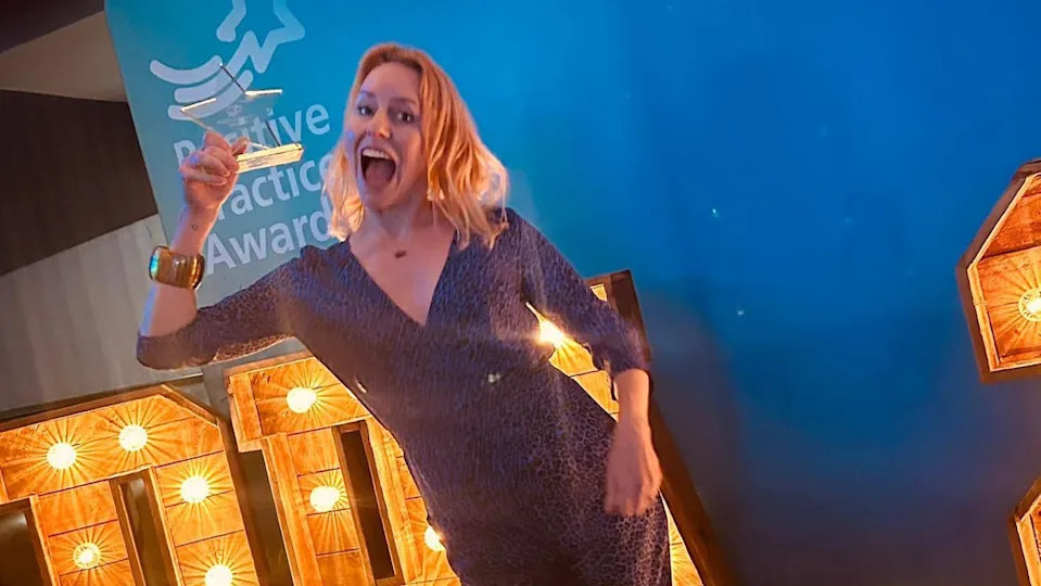 Natalie Rowlands wears a dark evening dress and holds up a trophy at an awards event, smiling into the camera.