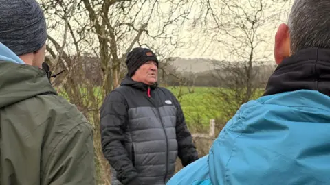 Martin Heath/BBC Mark O'Brien wearing a grey puffer jacket and black hat standing in front some bare trees and talking to a group of men. We see the backs of two of the men, one wearing a green jacket and the other a blue jacket.