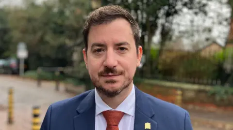 Sam Read/BBC Mike Reader stands outside the hospital. He is wearing a blue suit and orange tie. 