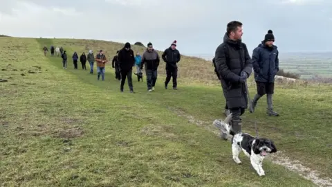 Martin Heath/BBC A line of men along with a couple of dogs, most dressed in anoraks and woollen hats, walking along a path on a grassy hillside with fields stretching off into the distance. The sky is cloudy.