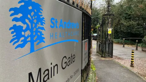 Sam Read/BBC Main gate of St Andrew's Healthcare. There is a metal sign on black railings, with blue logo shaped like a tree. To the right is an iron fence and some black and yellow striped bollards. 