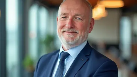 Steve Robertson Steve Robertson, a man with a close grey beard and blue eyes, wearing a blue suit and tie. He is looking into the camera with a slight smile