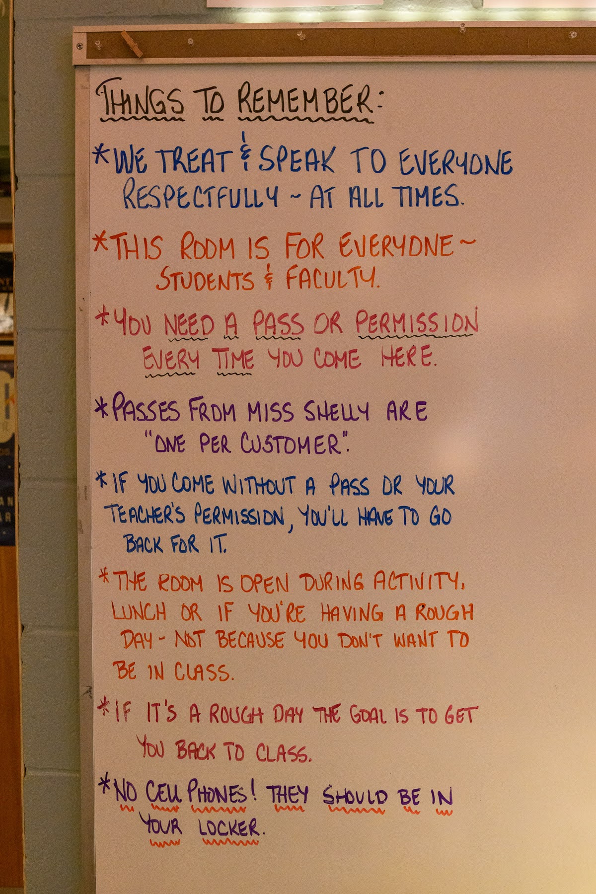 A whiteboard lists classroom rules about respect, entry permissions, needing passes, room access times, and a no cell phone policy, all written in multicolored markers.