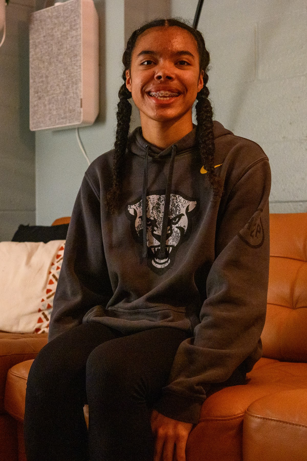 A person with braided hair and braces sits on a tan couch, wearing a dark hoodie with a graphic of a panther face and black pants.
