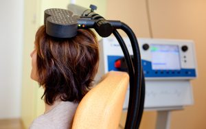 A woman sits in an exam chair with her side profile facing the camera and a medical device containing to flat black discs and hoses is perched on her head with a monitor in the background
