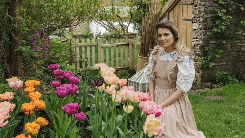 A woman wearing a brown frilly dress with a white puff sleeve shirt is kneeling in front of tulips in a garden. The tulips are all different colours, orange, purple and pink, and behind her is a lush garden.