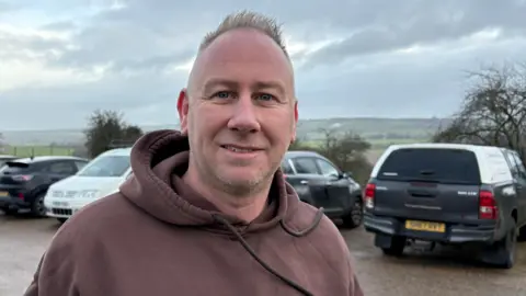Martin Heath/BBC Michael Cox with short grey hair, smiling at the camera and wearing a brown hoodie. He is standing in front of cars in a car park, with trees and fields behind.