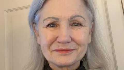 Handout Julie Colville is an older woman with long straight grey hair and blue eyes. He stands smiling in front of a cream-coloured door. 