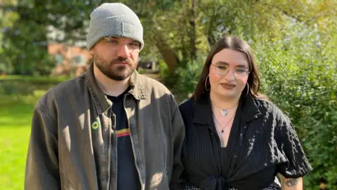 BBC A man on the left with glasses, a grey beanie, and a short brown beard is wearing a light brown shirt open over a dark t-shirt. A woman next to him on the right also has glasses and is dressed all in black with long brown hair. They are standing outside with trees in the background.