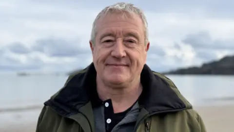BBC Jamie Dewar has short grey hair and is wearing a green outdoor jacket and a grey polo shirt underneath. He is standing on a beach with a calm see behind him and the shoreline running diagonally with the coastline visible to the right. It s a cloudy but fairly bright day.
