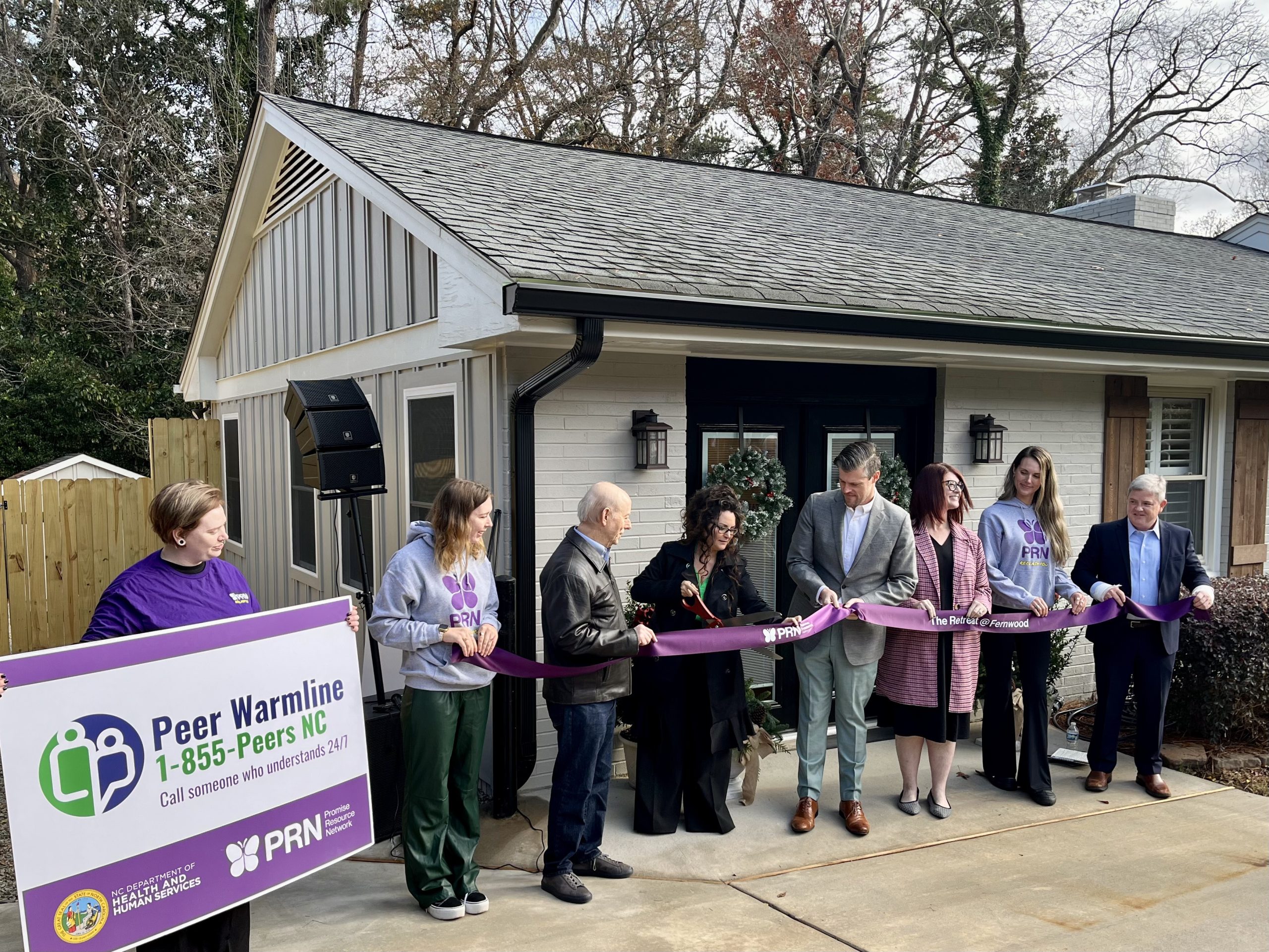 several people stand in front of a white painted brick ranch house holding a purple ribbon and the woman in the center is cutting it with giant scissors