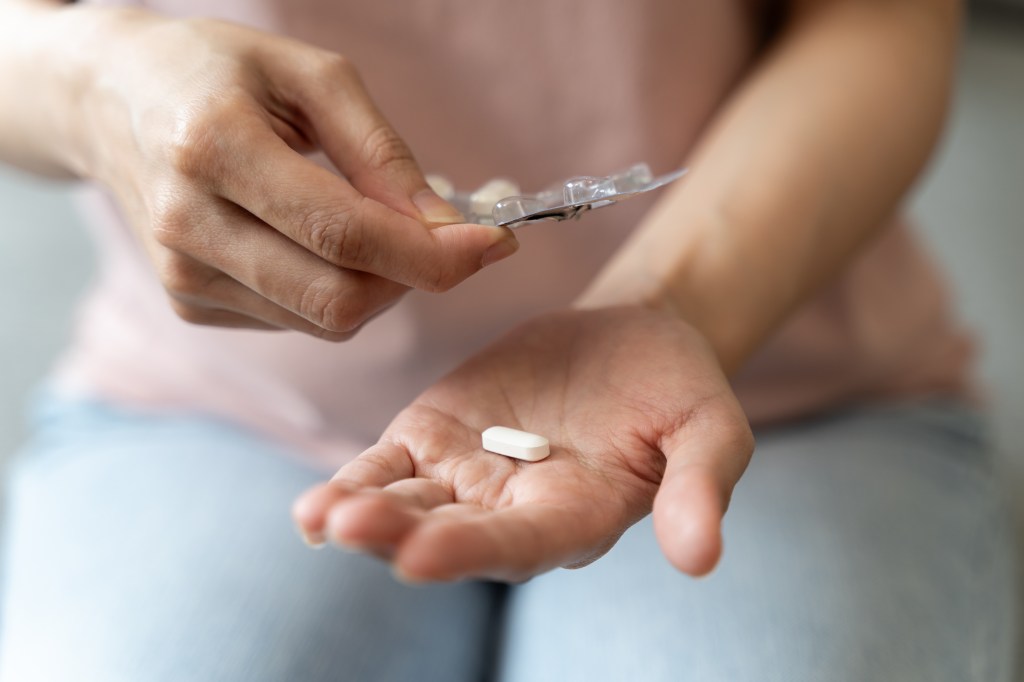 A hand holding a white rectangular pill in its palm, while the other hand squeezes another pill out of a blister pack.