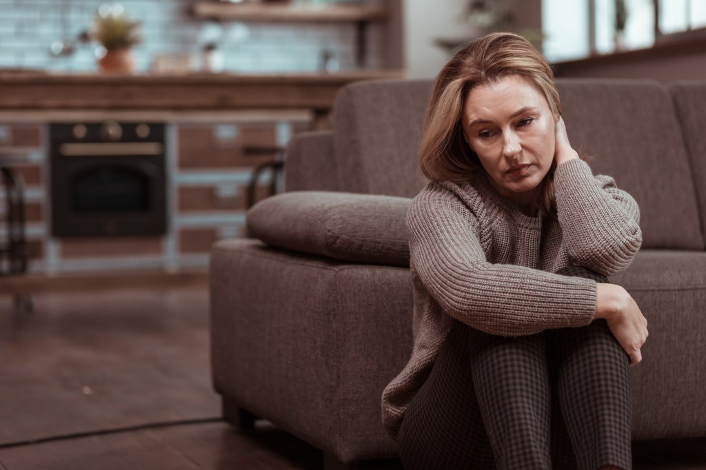 A woman wearing brown squared trousers feels depressed while sitting on the floor in her apartment.