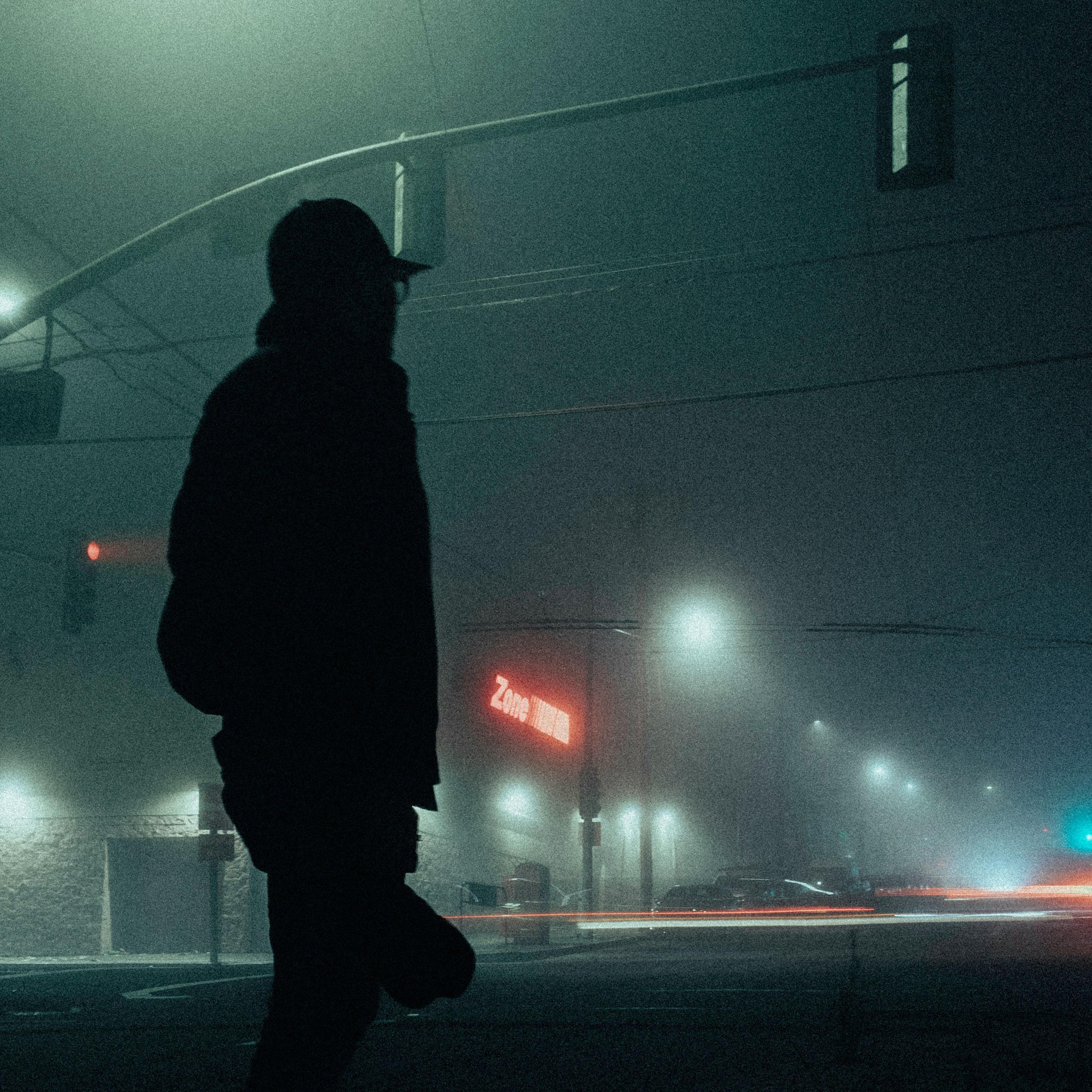 A silhouetted figure on a foggy street corner with traffic lights and blurred car lights at night.