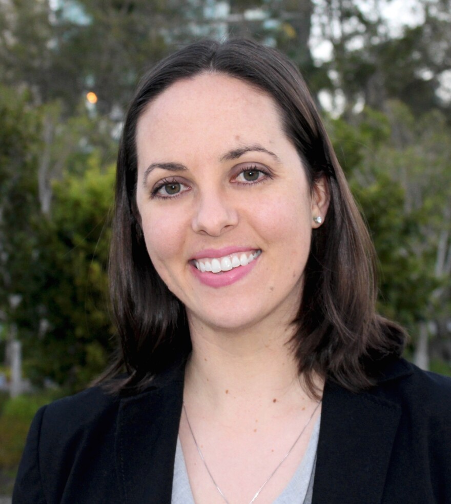A headshot photo of a smiling woman with brown hair and a black jacket.