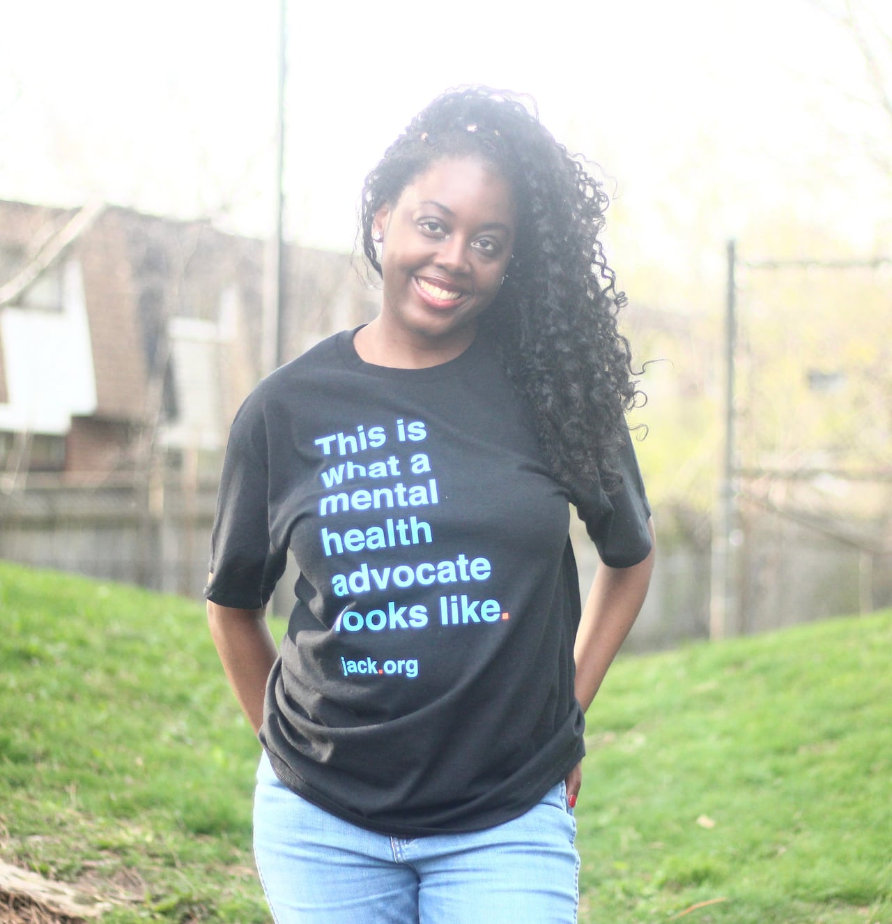 A smiling woman stands in a grassy field. She's wearing a shirt that says, "This is what a mental health advocate looks like."