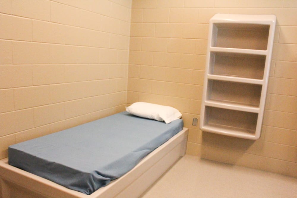 A room with a built in white set of shelves and a bed with a blue and mattress inside a state psychiatric hospital