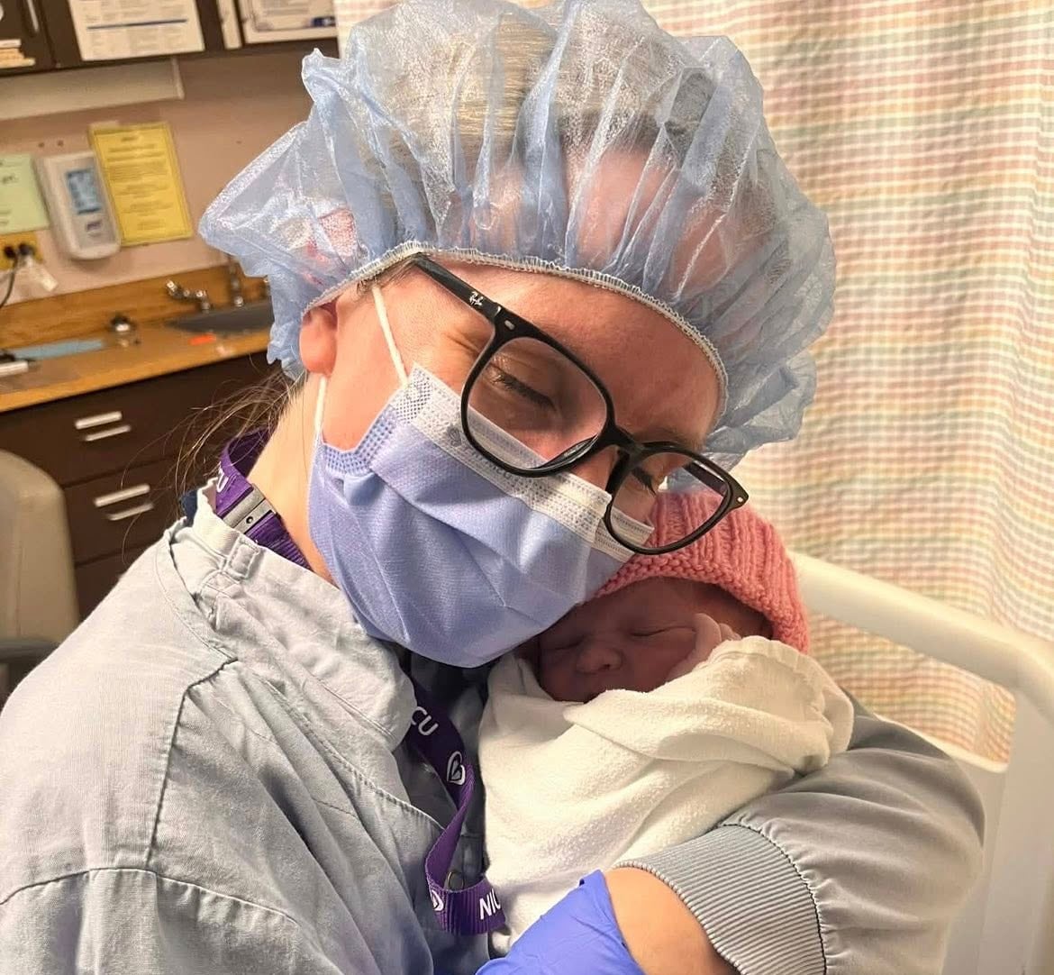 A nurse wearing a mask and gloves holding baby wrapped in a blanket.